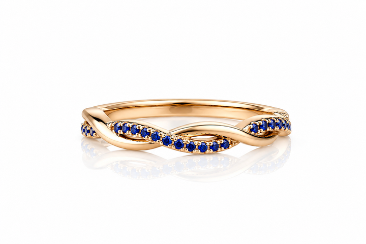 A twisted infinity-style ring in yellow gold, accented with rows of small round-cut deep blue sapphires on the front, shown on a clean white background with a soft reflection below.